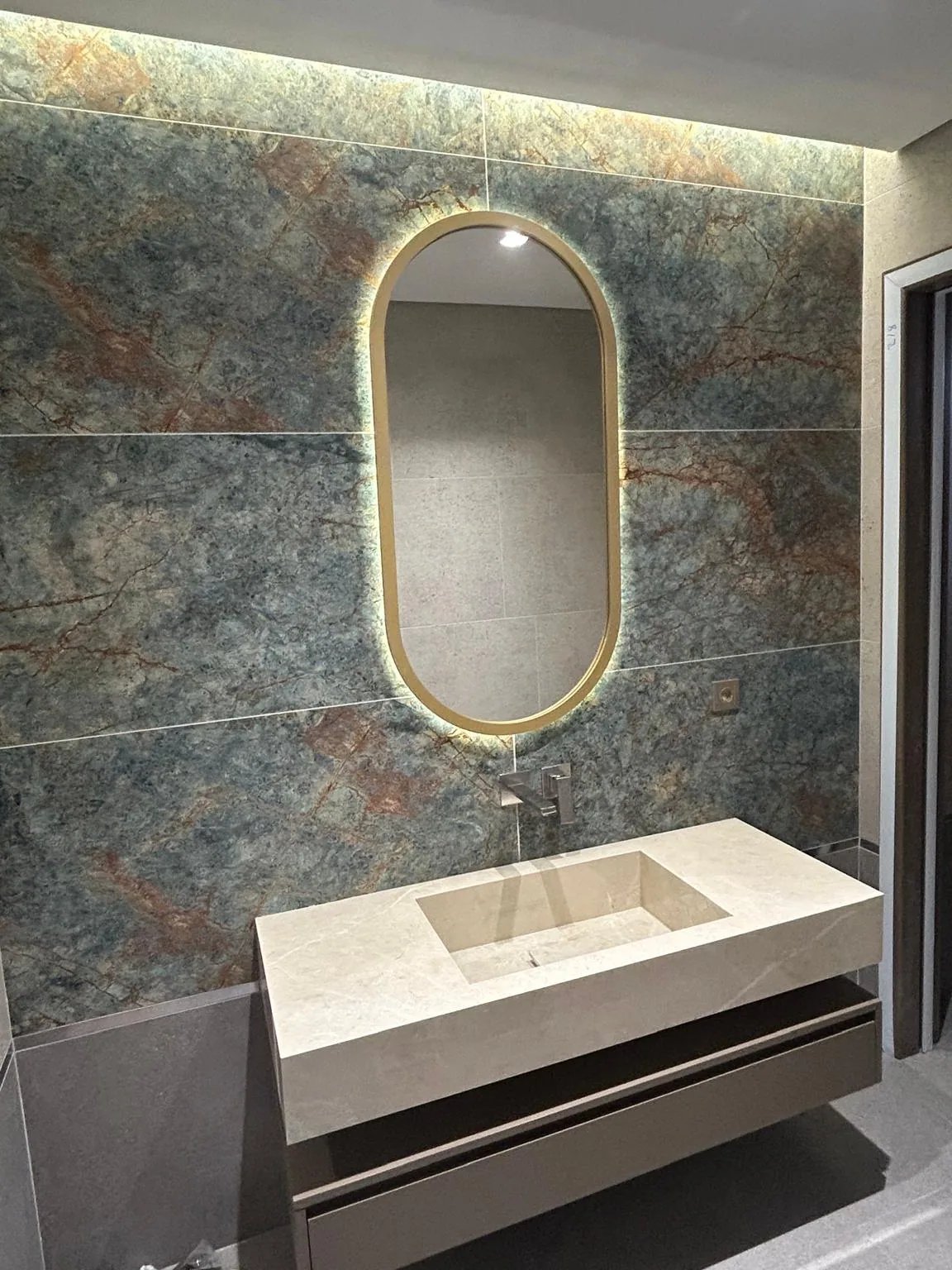 Bej mermer banyo tezgahı ve entegre lavabo teknesi; arkada yeşil damarlı mermer duvar kaplaması ve pirinç çerçeveli oval ayna