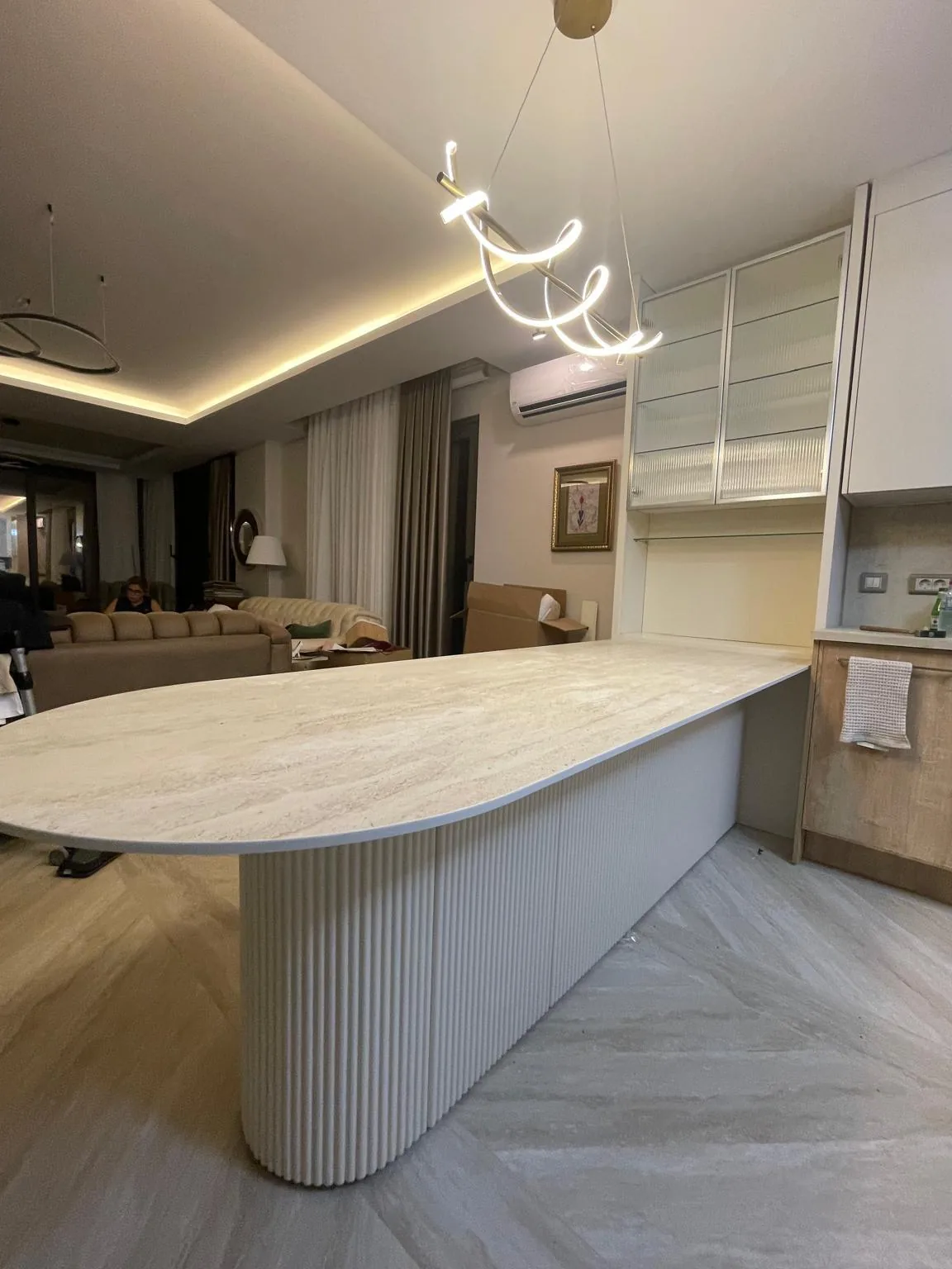 Yarım oval traverten mutfak adası — dikey yivli gövde ve kavis boyunca süren kalın kenar, açık plan salona bakıyor